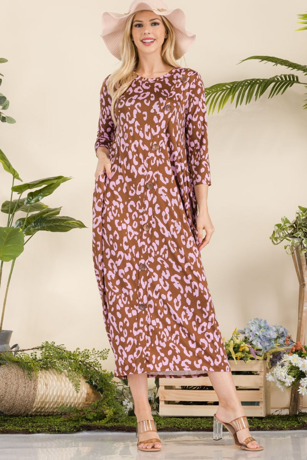 A woman in the Celeste Full Size Leopard Contrast Dress with pockets, featuring a brown and pink leopard print design and 3/4 sleeves, stands smiling among potted plants and wooden crates while wearing a wide-brim hat and sandals.