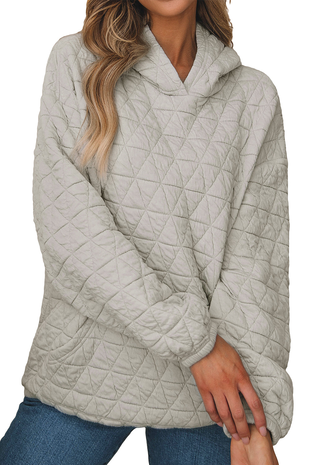 A woman stands partially turned away, wearing a Light Grey Solid Color Quilted Kangaroo Pocket Hoodie with a drop shoulder design and blue jeans. She touches her hair with her right hand, and there are beige and brown decor elements in the background.