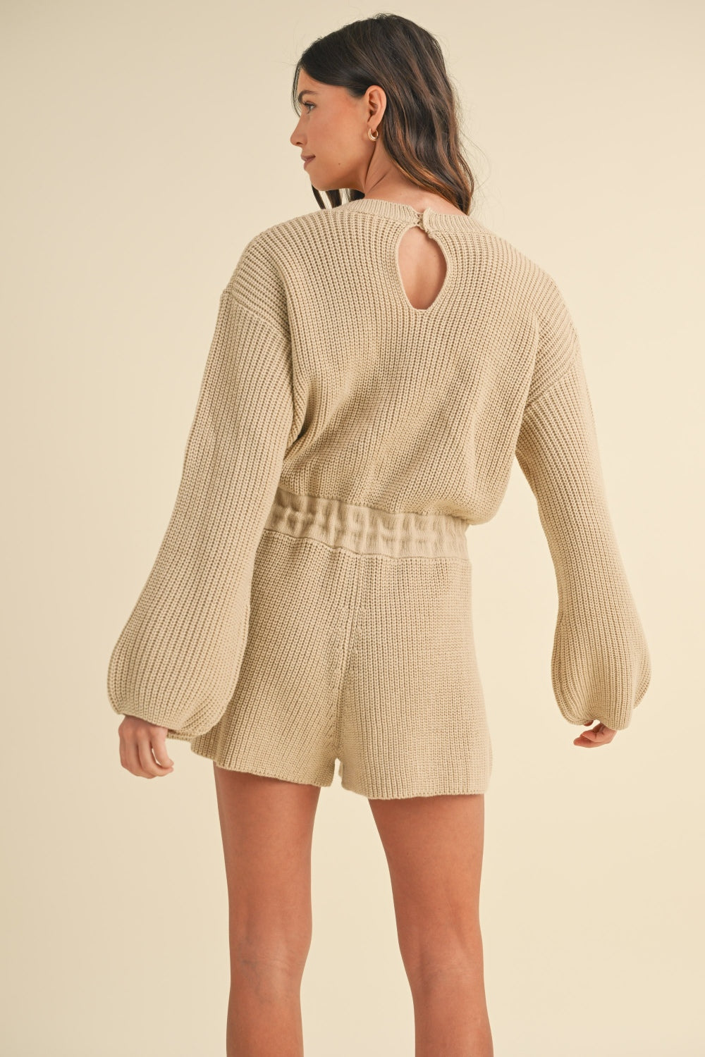 A woman is wearing the MABLE Round Neck Long Sleeve Sweater Romper in a cozy beige knit, complete with matching shorts. With one hand casually touching her hair, she stands against a plain background, effortlessly exuding style.