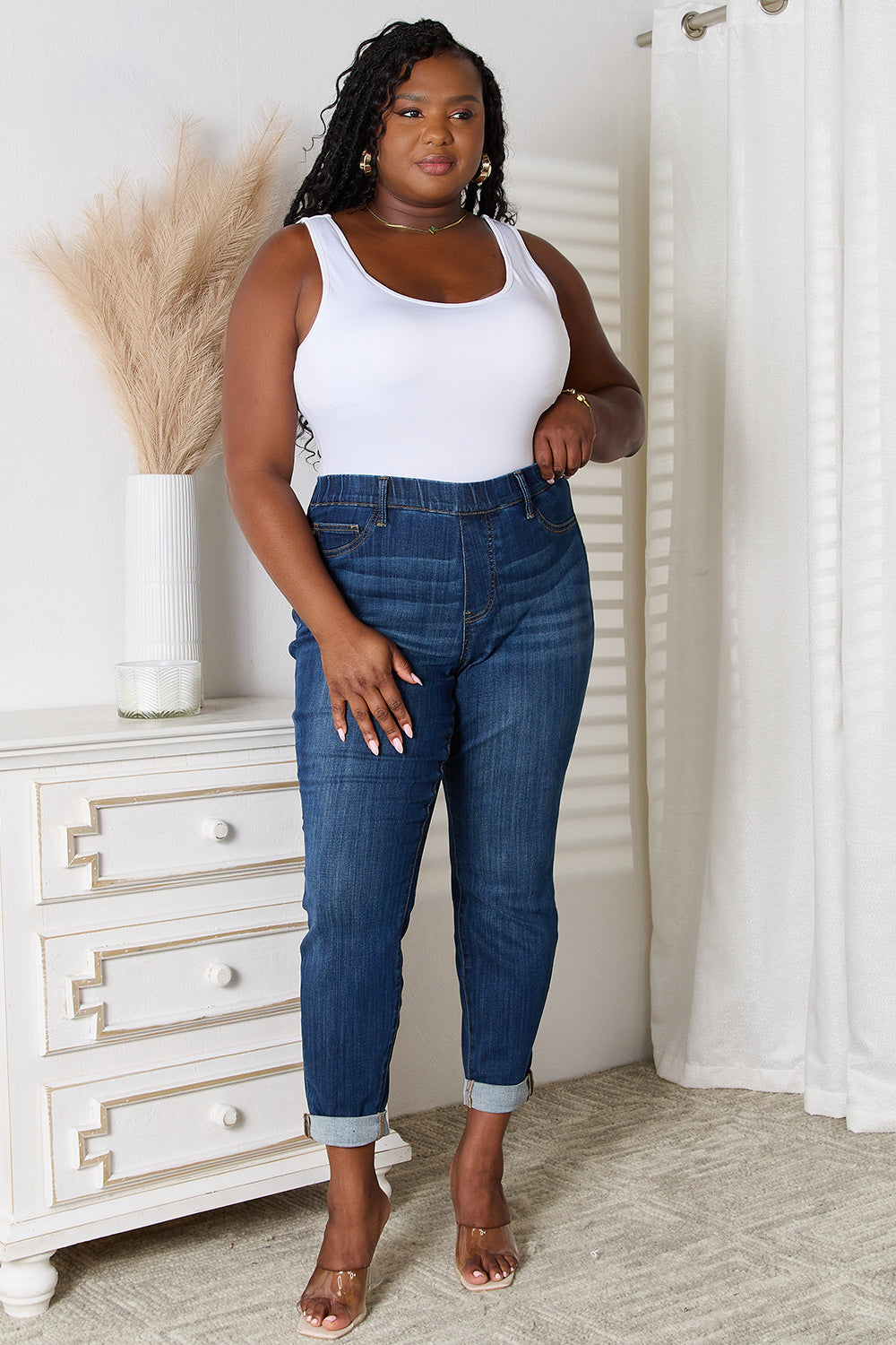 A person wearing Judy Blue Full Size Skinny Cropped Jeans with rolled-up cuffs and clear heels stands near a white dresser adorned with a decorative vase for a modern look.