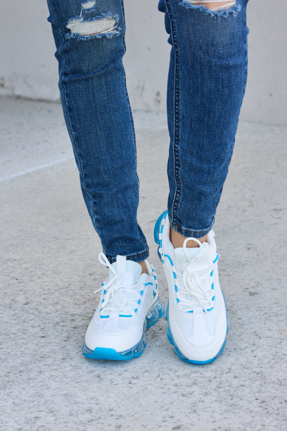 A person wearing ripped jeans and stylish Forever Link Lace-Up Air-Cushioned Athletic Shoes in white with blue accents stands confidently on a concrete surface.