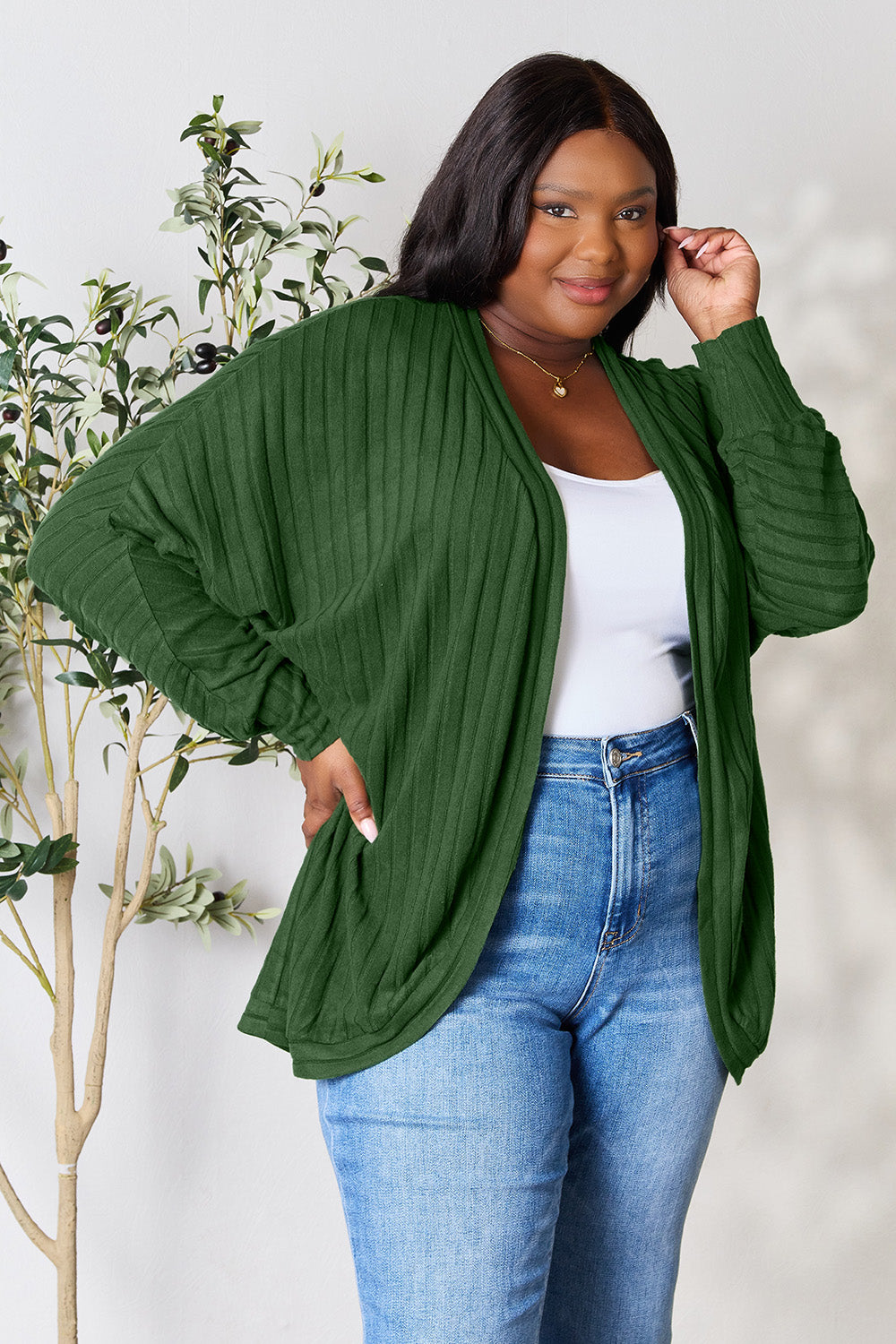 A person with wavy hair, wearing the Basic Bae Full Size Ribbed Cocoon Cardigan over a white top and beige pants, stands in front of a light-colored background with a leafy plant, showcasing an effortlessly basic style.