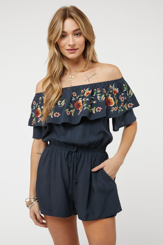 Wearing the stylish Floral Embroidered Off Shoulder Romper, a woman poses gracefully, showcasing its ruffle detail and making it an ideal addition to any summer wardrobe.