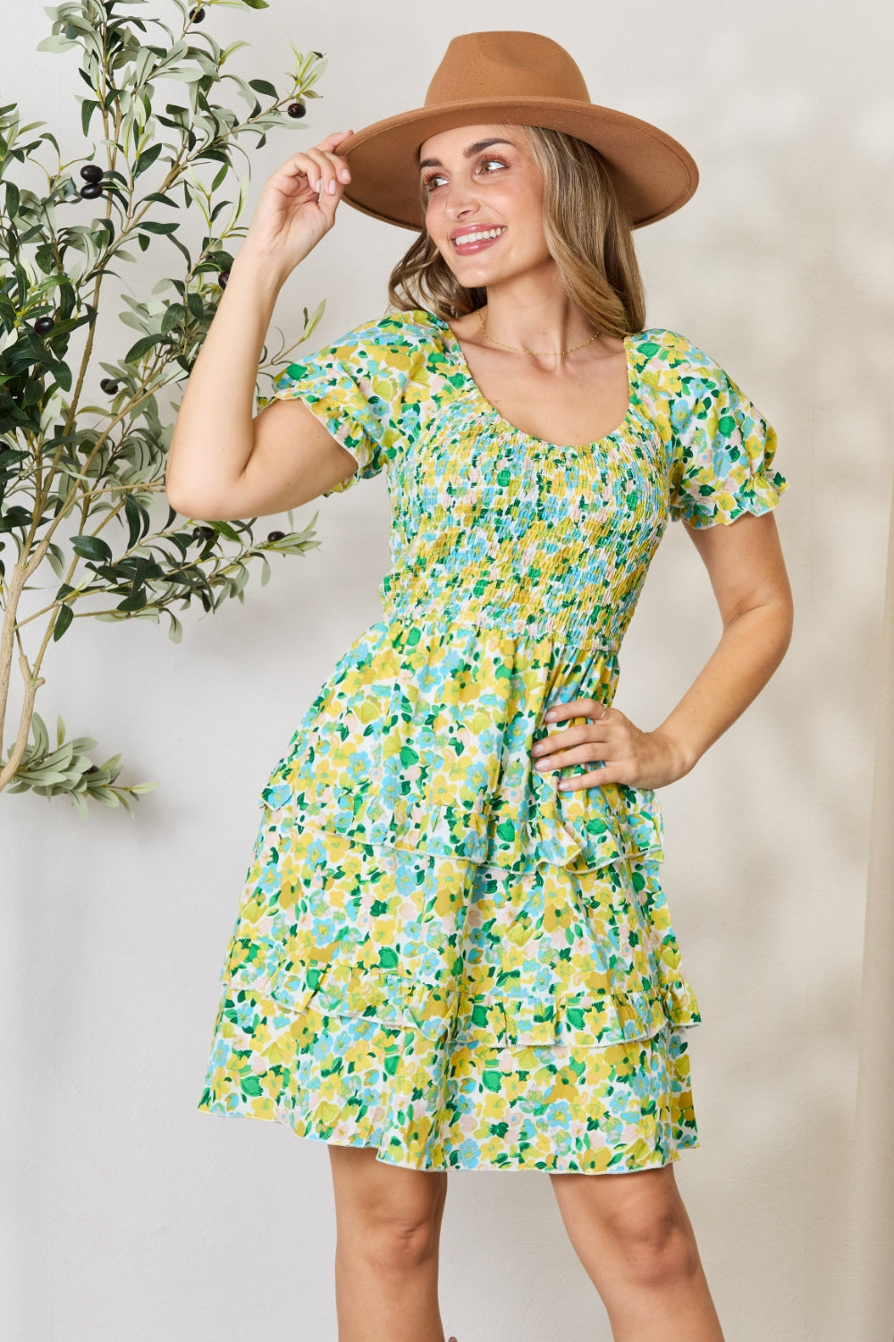 A woman wearing the Devine Smocked Scoop Neck Flounce Sleeve Mini Dress and a brown hat smiles and poses near greenery indoors.