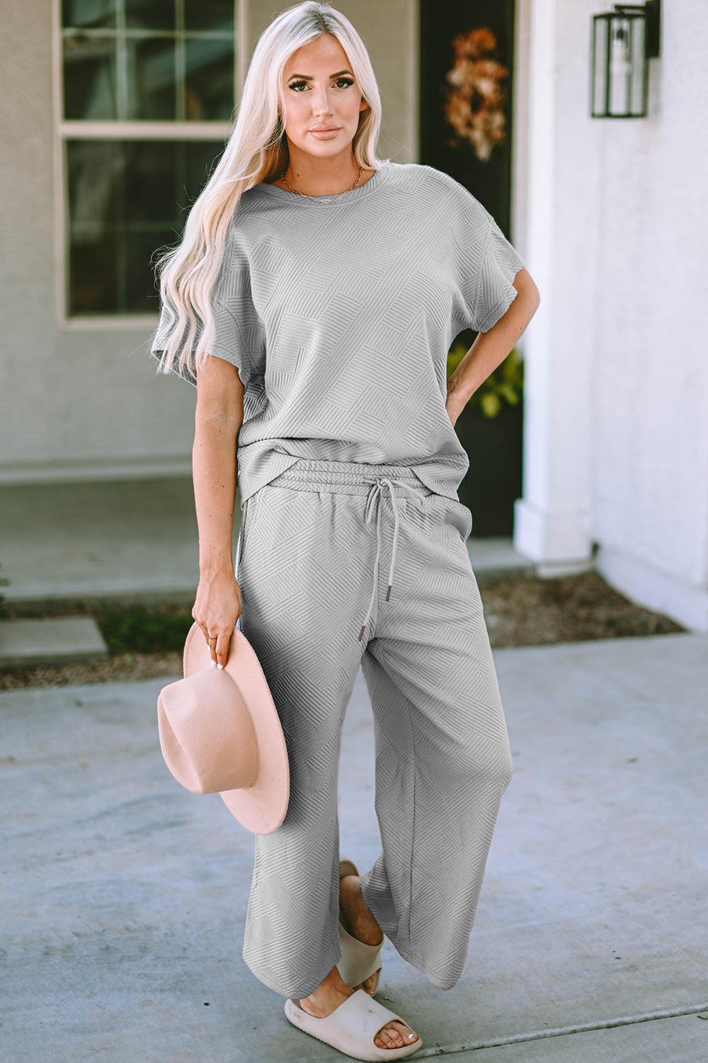 A woman with long hair smiles while standing outside a building, dressed in the Double Take Full Size Texture Short Sleeve Top and Pants Set, featuring a matching beige two-piece ensemble. She wears slides and holds a hat in her right hand. Her outfit, slightly stretchy for added comfort, is perfect for machine wash cold.