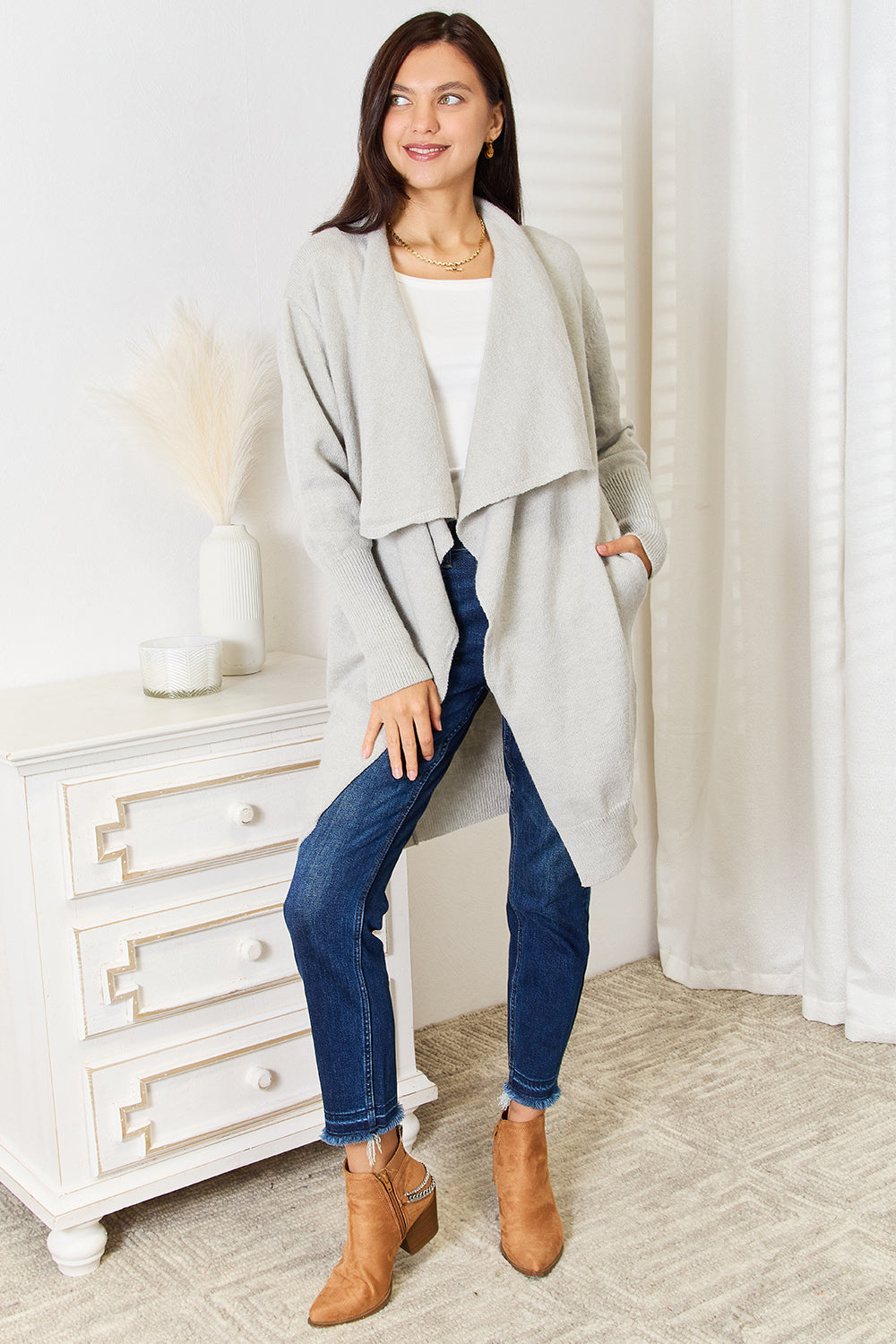 A person with long brown hair wearing a gray Angel Wings Open Front Duster Cardigan with Pockets and blue jeans stands next to a white dresser with a vase and candle on it.