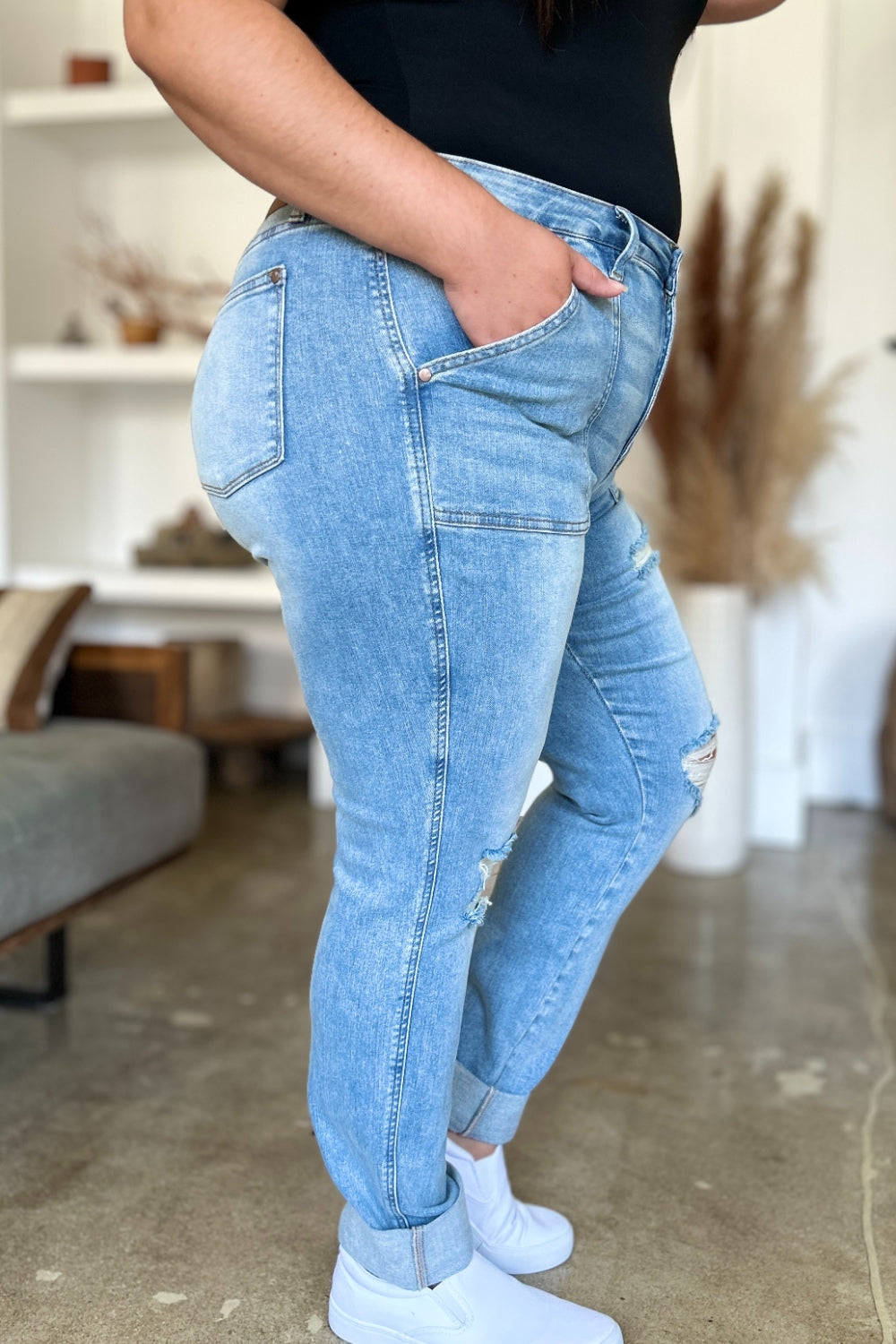 Two women are standing side by side, both clad in black and white tank tops and Judy Blue Full Size Distressed Straight Jeans with Patch Pockets, smiling at the camera in a modern living room.