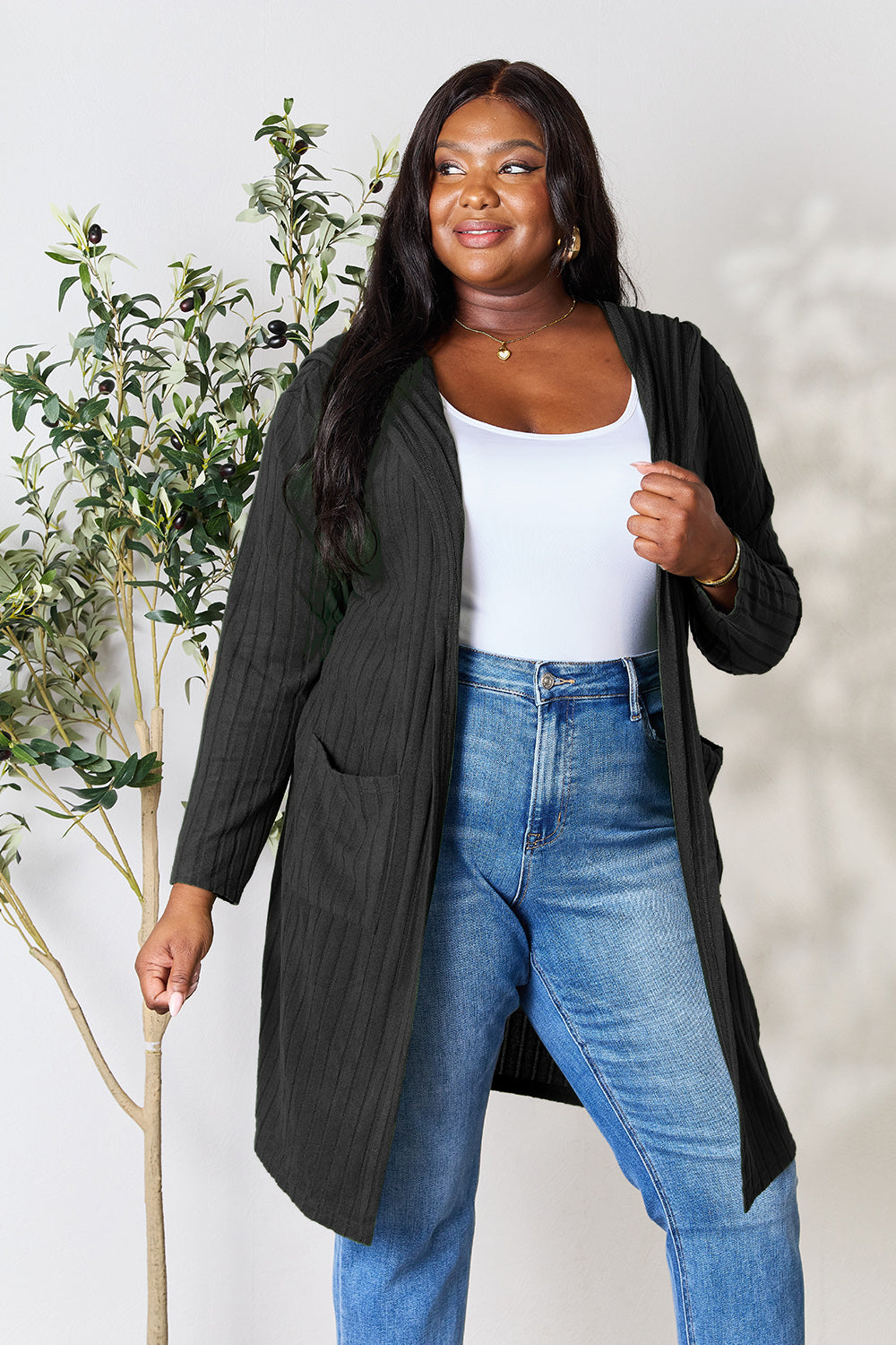 A woman wearing the Basic Bae Full Size Hooded Sweater Cardigan in black, paired with a white top and khaki pants, stands in front of a light background accented by a leafy plant on her left. With one hand tucked into her pocket, she gazes to her right.