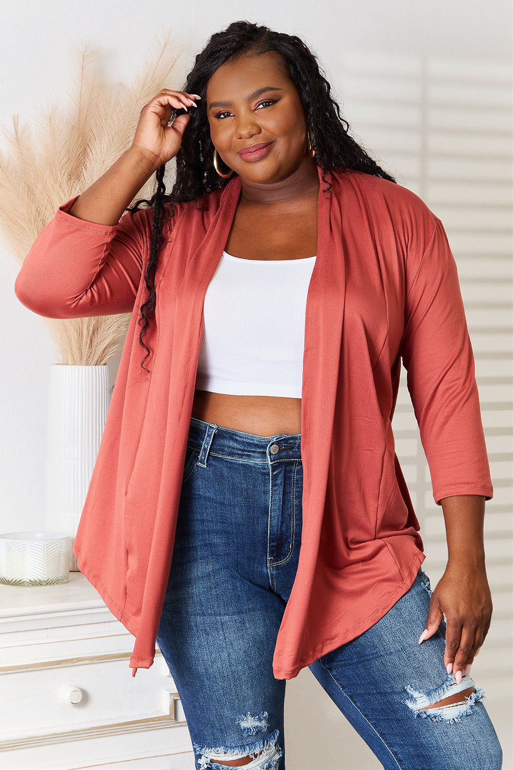 A person with long hair is wearing the Culture Code Full Size Open Front Cardigan in a rust color, complemented by a flattering draped front. They pair it with a white top and ripped jeans while standing indoors with one hand on their hip.