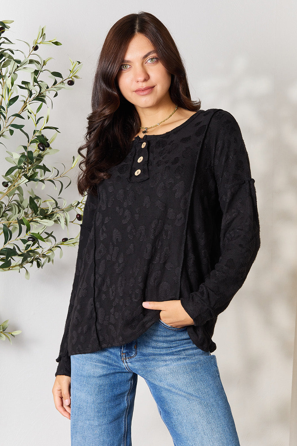 A woman wearing a relaxed fit, black BOMBOM Textured Exposed Seam Buttoned Blouse and jeans stands with one hand in her pocket next to a leafy plant.