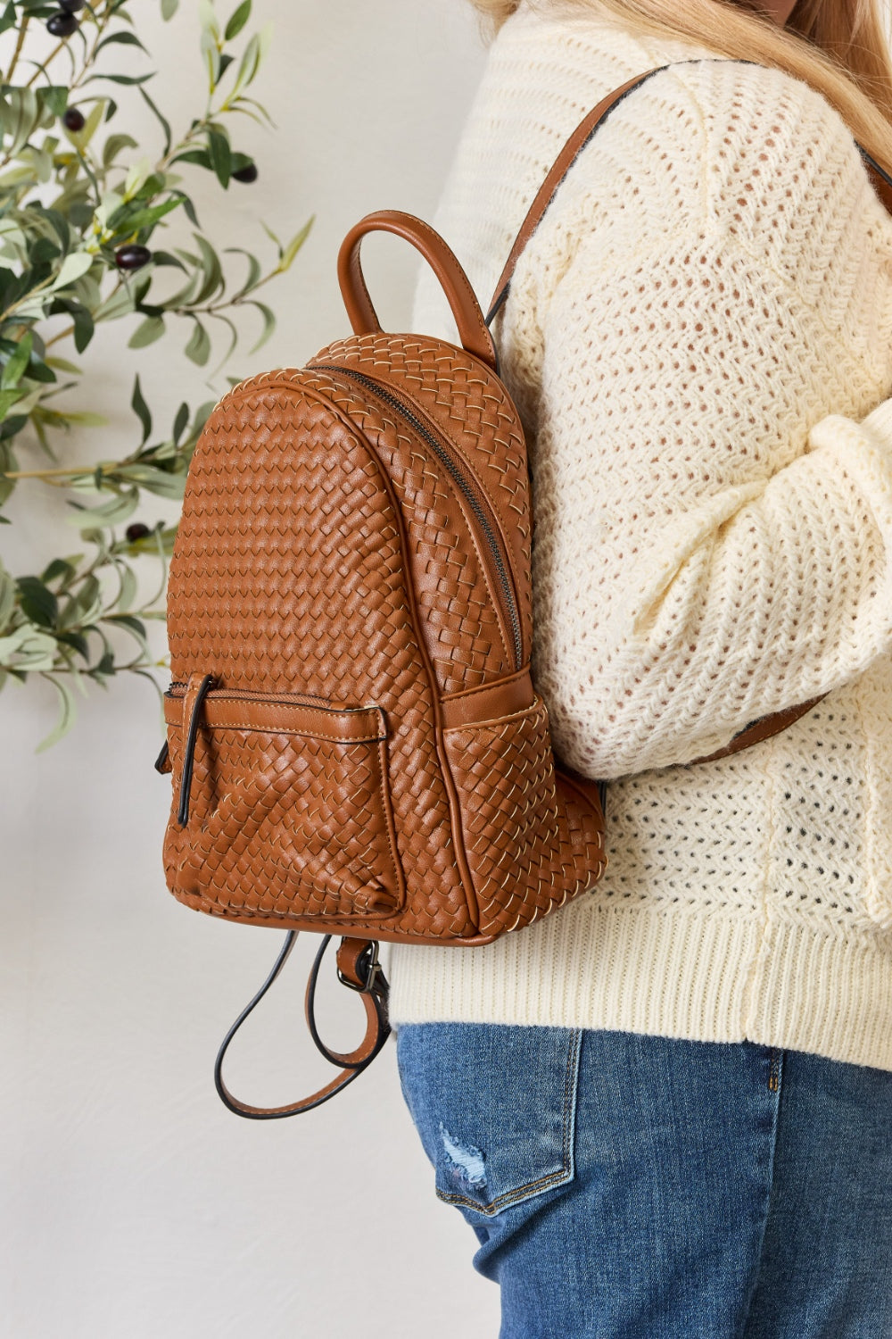 A person wearing a cream sweater and jeans is carrying the SHOMICO PU Leather Woven Backpack in brown.