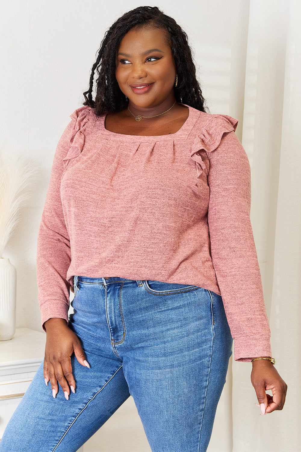 A woman wearing a Double Take Square Neck Ruffle Shoulder Long Sleeve T-Shirt in pink and blue jeans stands with one hand in her pocket, smiling. She has long curly hair and wears a necklace and bracelet. She stands in a room with white decor.