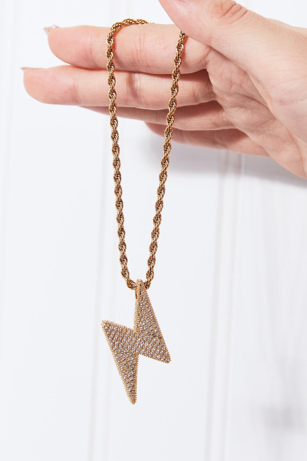 A Brass Lightning Pendant Necklace, featuring a gold twisted chain and a lightning bolt-shaped pendant encrusted with small cubic zirconia stones, displayed on a light blue background.