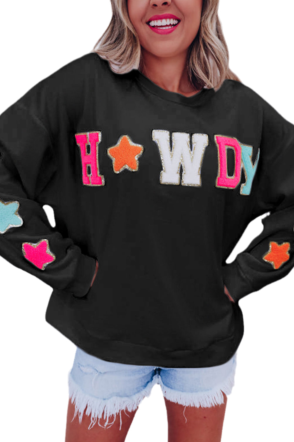 A woman with short blonde hair is wearing the Black Howdy Glitter Chenille Patch Graphic Casual Sweatshirt, featuring colorful star patterns and "WD" letters. She pairs it with light denim shorts and smiles while looking to the side.