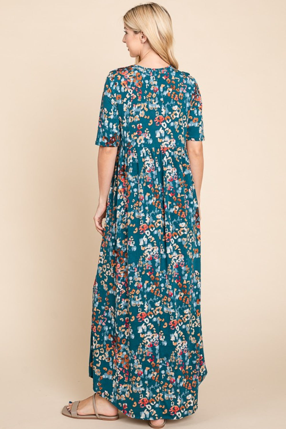 A person wearing a BOMBOM Printed Shirred Maxi Dress, featuring an eye-catching teal floral pattern and short sleeves, stands against a beige background.