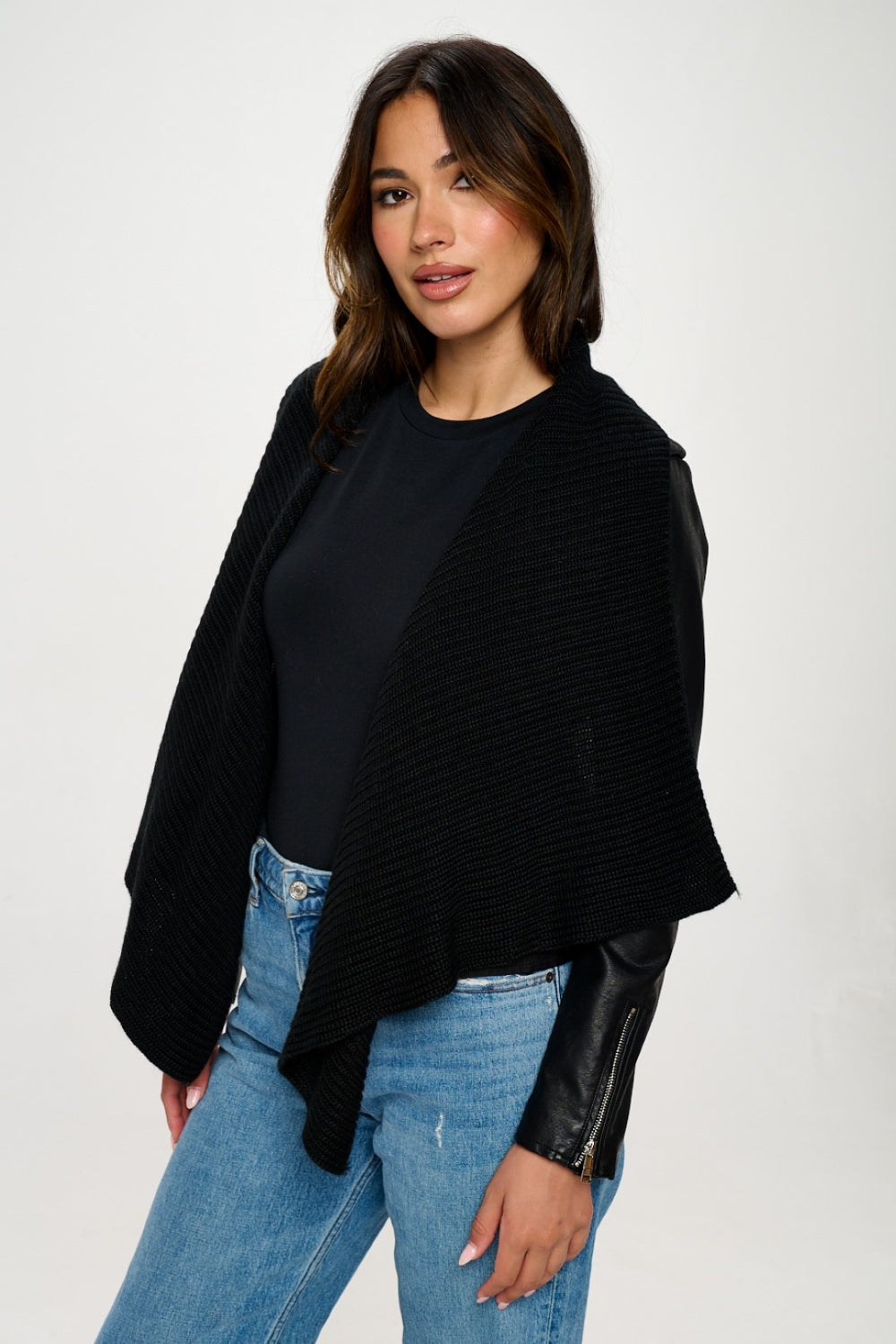 A woman with long, dark hair wearing a black top, a black shawl over the Coalition LA Knit Collared Faux Leather Crop Jacket, and blue jeans stands against a plain white background.