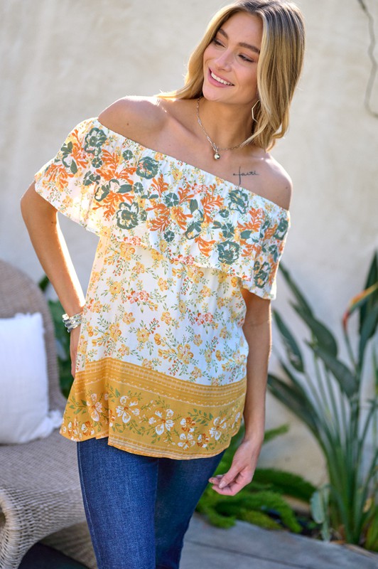 A woman with blonde hair is smiling while standing outdoors, wearing the Printed Off Shoulder Smocked Top paired with jeans.