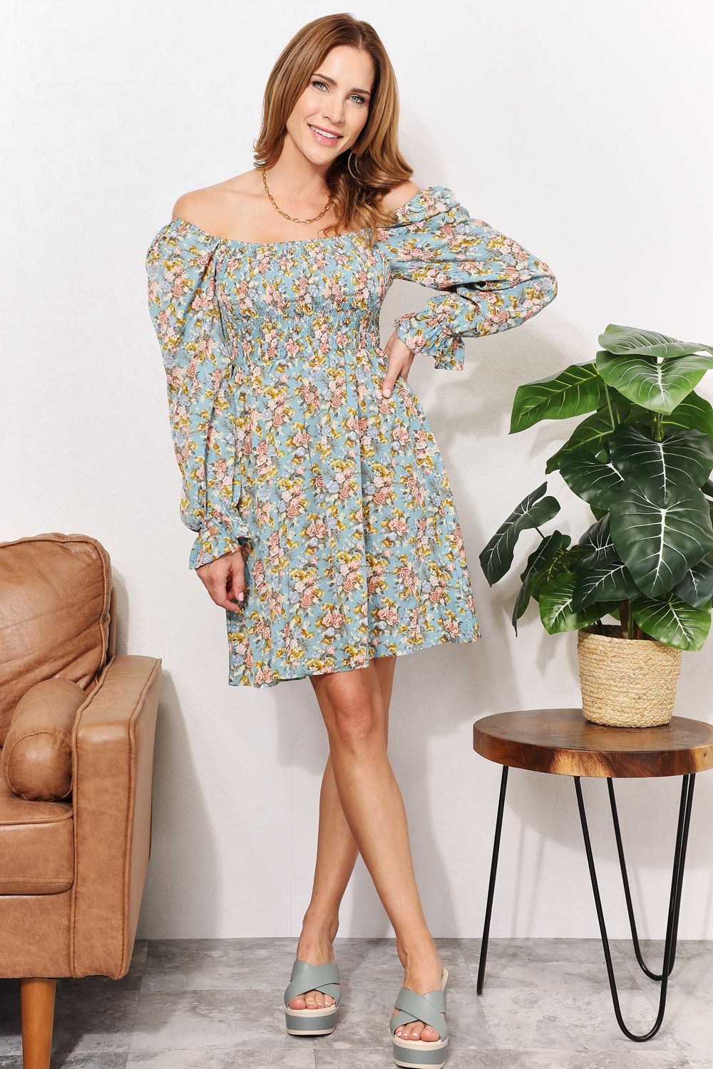 A woman in the Honey Floral Smocked Flounce Sleeve Square Neck Dress stands indoors, holding the hem of her dress. She is smiling slightly with her eyes looking downward. Nearby, a plant and a brown couch complete the cozy scene.