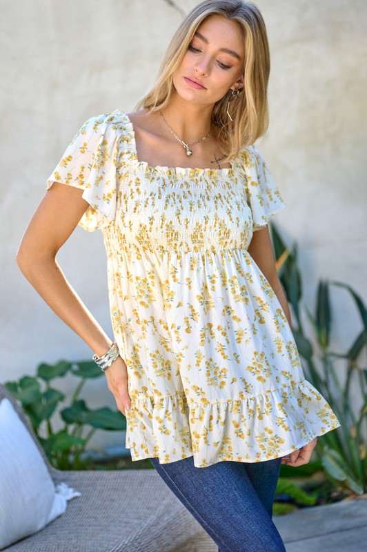 A woman stands outdoors donning a Floral Printed V-Neck Ruffle Top paired with jeans, set against a neutral background, with plants in the foreground, highlighting her fashion-forward wardrobe.