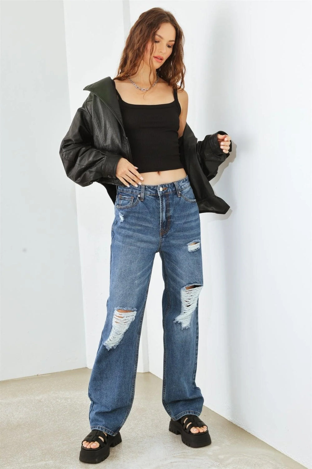 Person wearing HAMMER COLLECTION Distressed High Waist Jeans with ripped knees and black platform sandals, standing in a minimalist setting. The flattering fit and edgy distressed details add to the overall chic look.