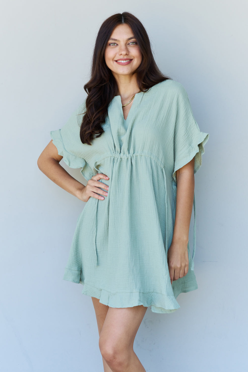 A woman is wearing the Ninexis Out Of Time Full Size Ruffle Hem Dress with Drawstring Waistband in Light Sage. Her long, dark hair falls gracefully as she smiles and looks to her left, standing against a plain background.