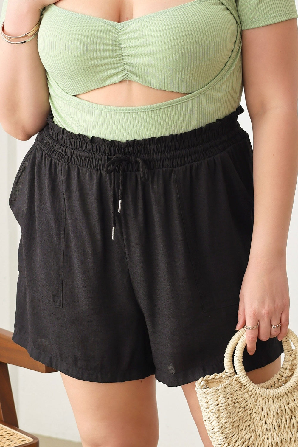 A woman wearing a light green cut-out top and Zenobia Plus Size Drawstring Elastic Waist Shorts with Pockets stands indoors, holding a woven handbag.