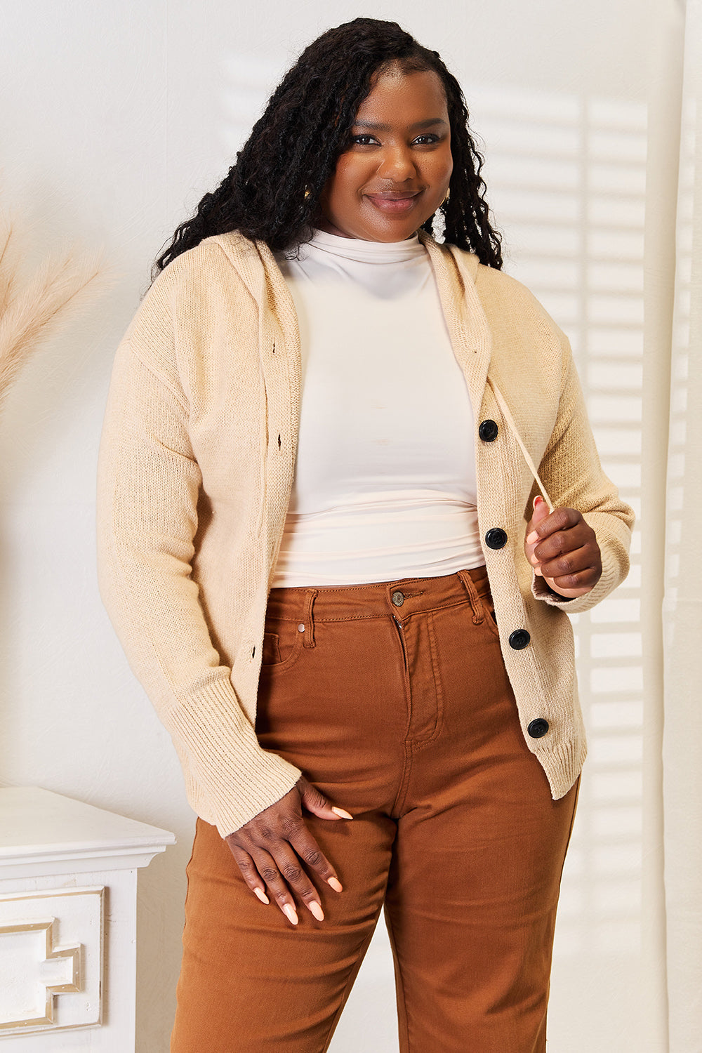 A person stands in a well-lit room wearing the Woven Right Button-Down Long Sleeve Hooded Sweater in a cream color over a white top and brown pants. The individual smiles slightly and poses with one hand in a pocket, against a light background.