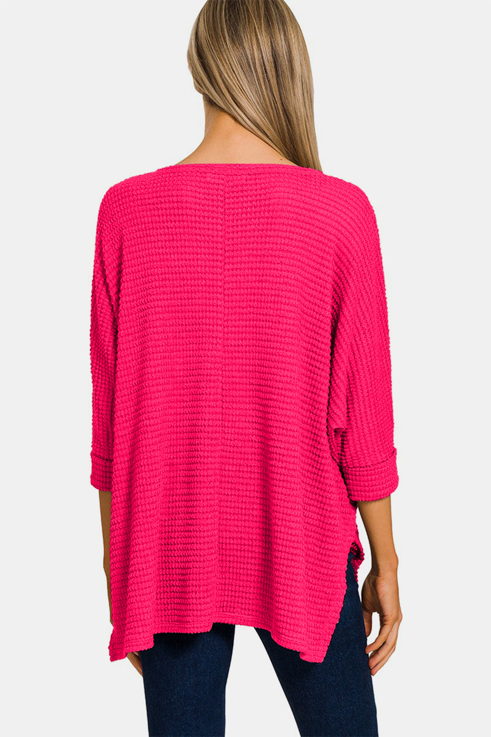 A woman wearing the Zenana V-Neck High-Low Jacquard Knit Top in a bright pink, loose-knit style with three-quarter sleeves, paired with blue jeans, stands against a plain white background, showcasing her comfortable knit top.