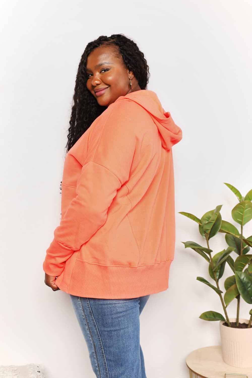 A woman with long hair wears a Double Take Quarter-Snap Dropped Shoulder Hoodie in solid orange. She is standing, with one hand touching her hair and the other resting in the pocket of her jeans, exuding a casual vibe.