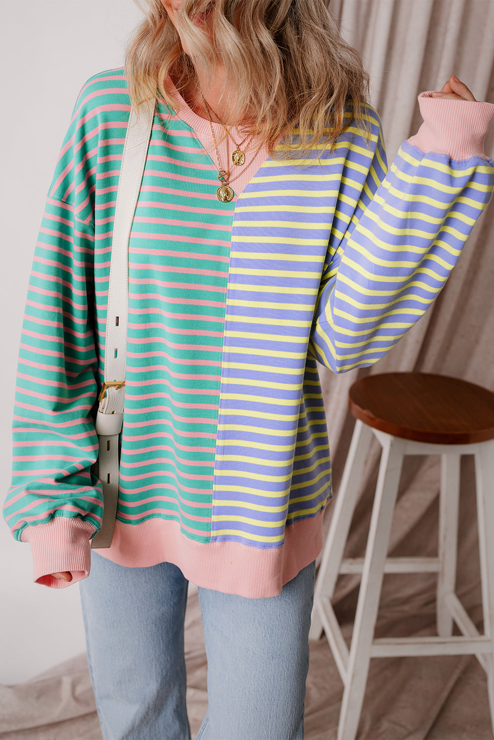 A person with wavy hair is seen from behind wearing a Casual Stripe Colorblock Drop Shoulder Oversize Sweatshirt in pastel colors and blue jeans. They are carrying a white shoulder bag, showcasing a trendy color block design.