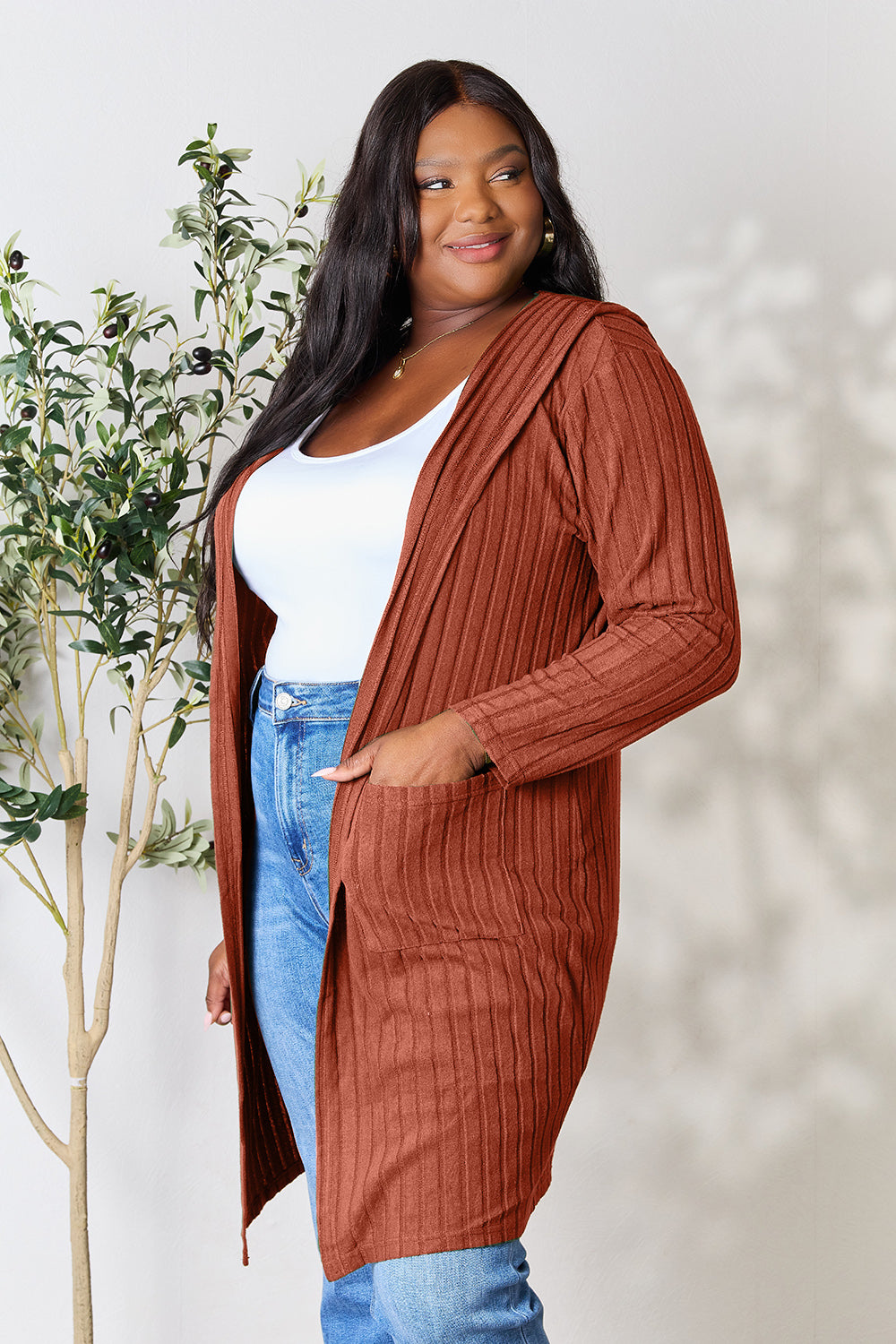 A woman wearing the Basic Bae Full Size Hooded Sweater Cardigan in black, paired with a white top and khaki pants, stands in front of a light background accented by a leafy plant on her left. With one hand tucked into her pocket, she gazes to her right.