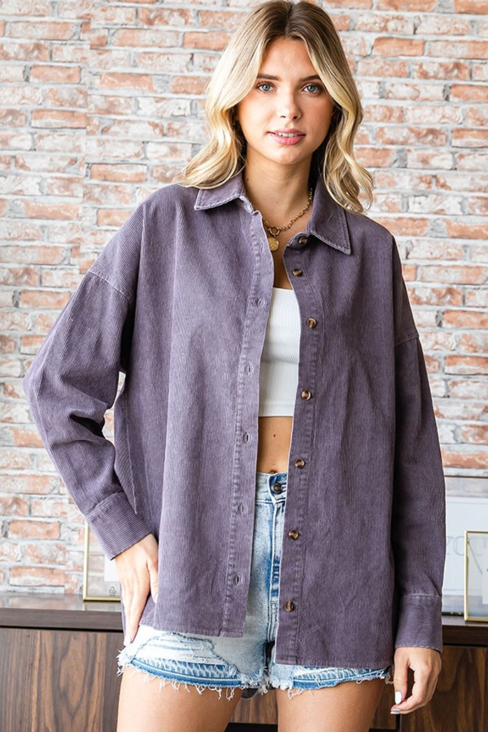 A blonde woman wearing the First Love Washed Corduroy Button Down Long Sleeve Shirt in purple over a white crop top and denim shorts stands in front of a brick wall.