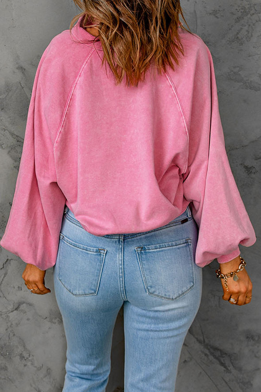 A person with wavy brown hair, wearing a loose-fit Pink Washed Snap Buttons Lantern Sleeve Pullover Sweatshirt and light blue jeans, is standing with their back facing the camera.