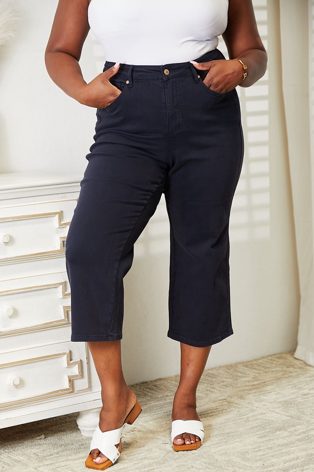 A person wearing a white sleeveless top and Judy Blue Full Size High Waist Tummy Control garment-dyed wide cropped jeans in black stands indoors next to a white dresser.