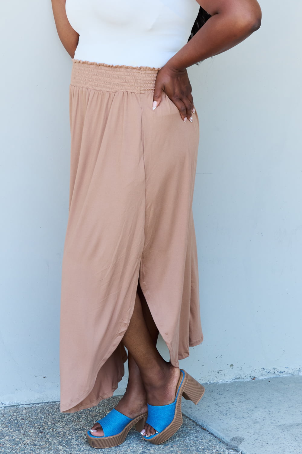 A person stands against a wall, wearing a white sleeveless top, the Doublju Comfort Princess Full Size High Waist Scoop Hem Maxi Skirt in Tan, and blue platform sandals. The right hand rests on the side of the skirt.