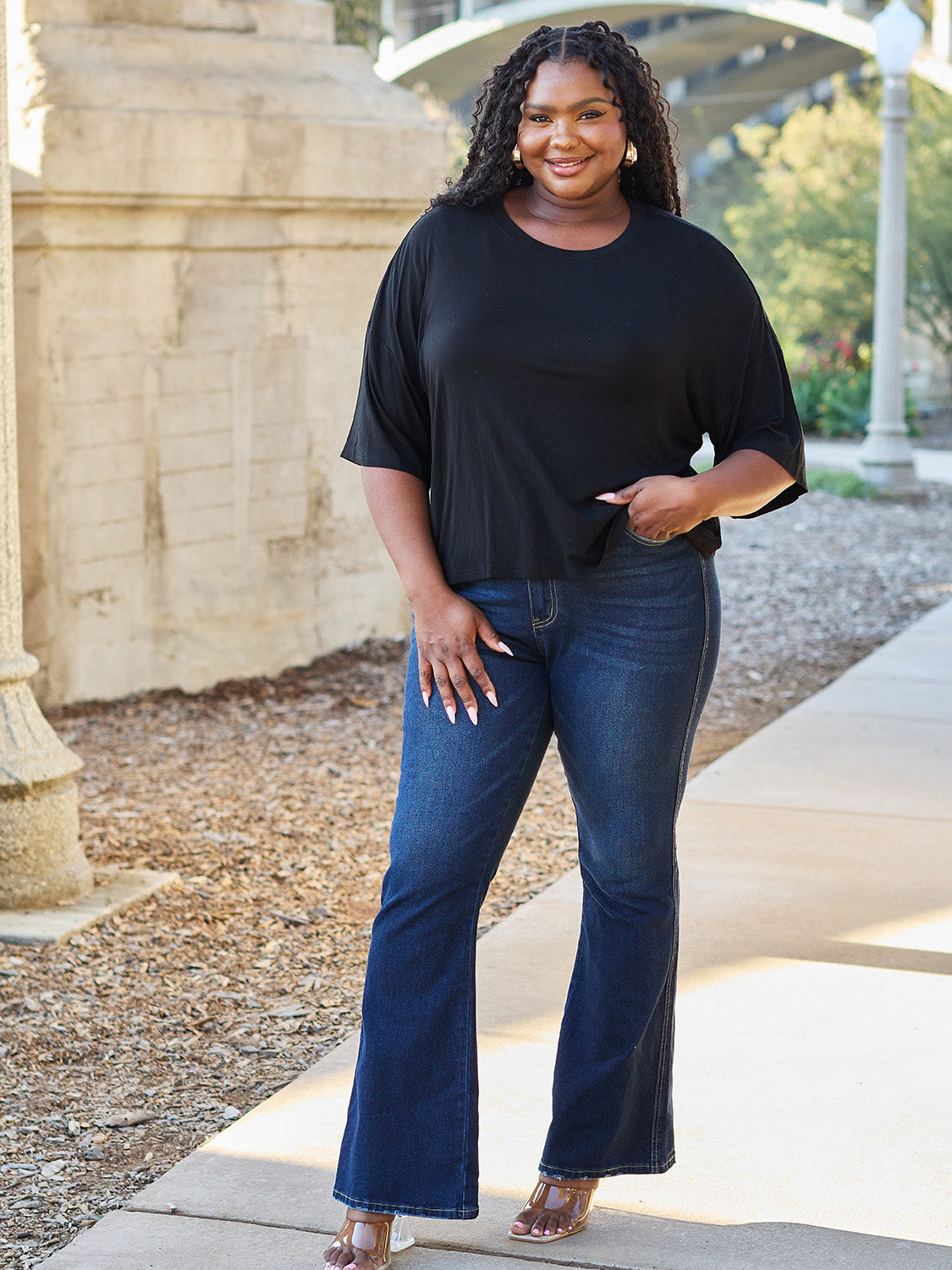 A person with long dark hair and a bright smile stands outdoors, showcasing their basic style in a loose, slightly stretchy *Basic Bae Full Size Round Neck Drop Shoulder T-Shirt* in pink and blue jeans, with one hand on their hip.