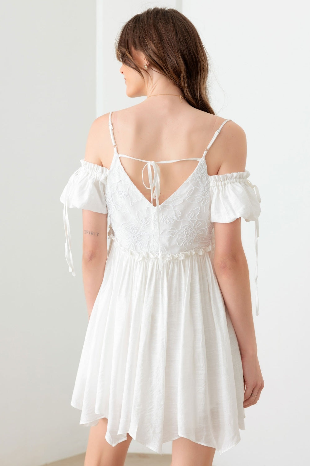 A woman poses in a boho-inspired, embroidered floral shirred cold shoulder mini dress by Mustard Seed. She accessorizes with a gold necklace and maintains a neutral expression against the backdrop of a plain white wall.