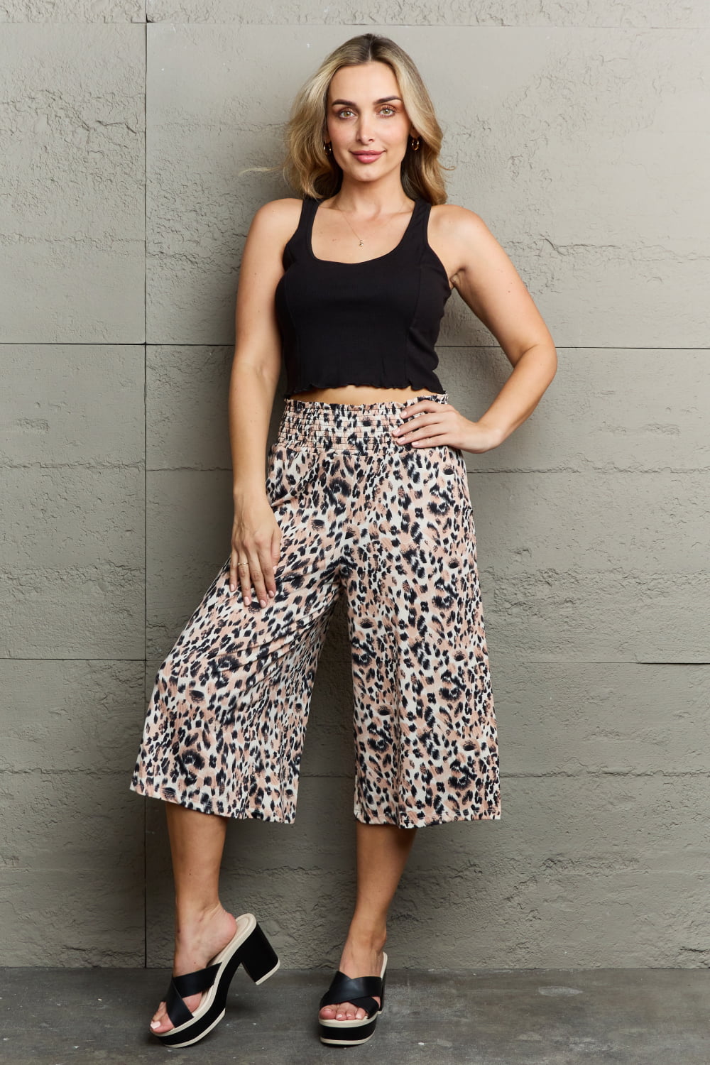 A person standing against a wall, wearing a black sleeveless top, chic Ninexis Leopard High Waist Flowy Wide Leg Pants with Pockets, and black platform sandals. One hand is in their pocket.