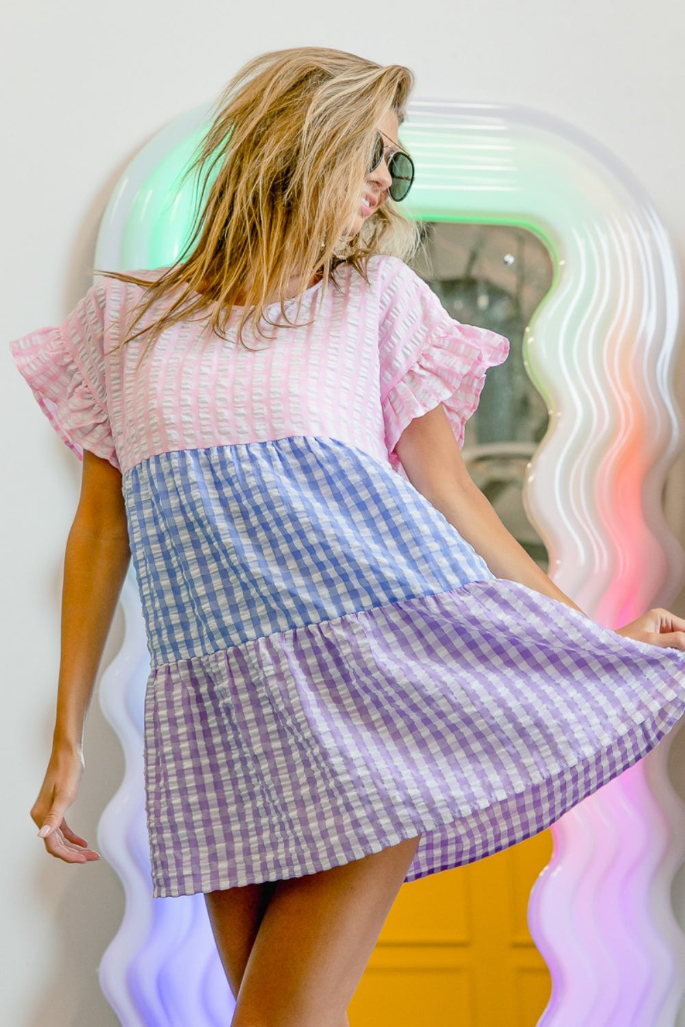 A person in sunglasses and the BiBi Plaid Color Block Tiered Top poses in front of a wavy, colorful mirror.