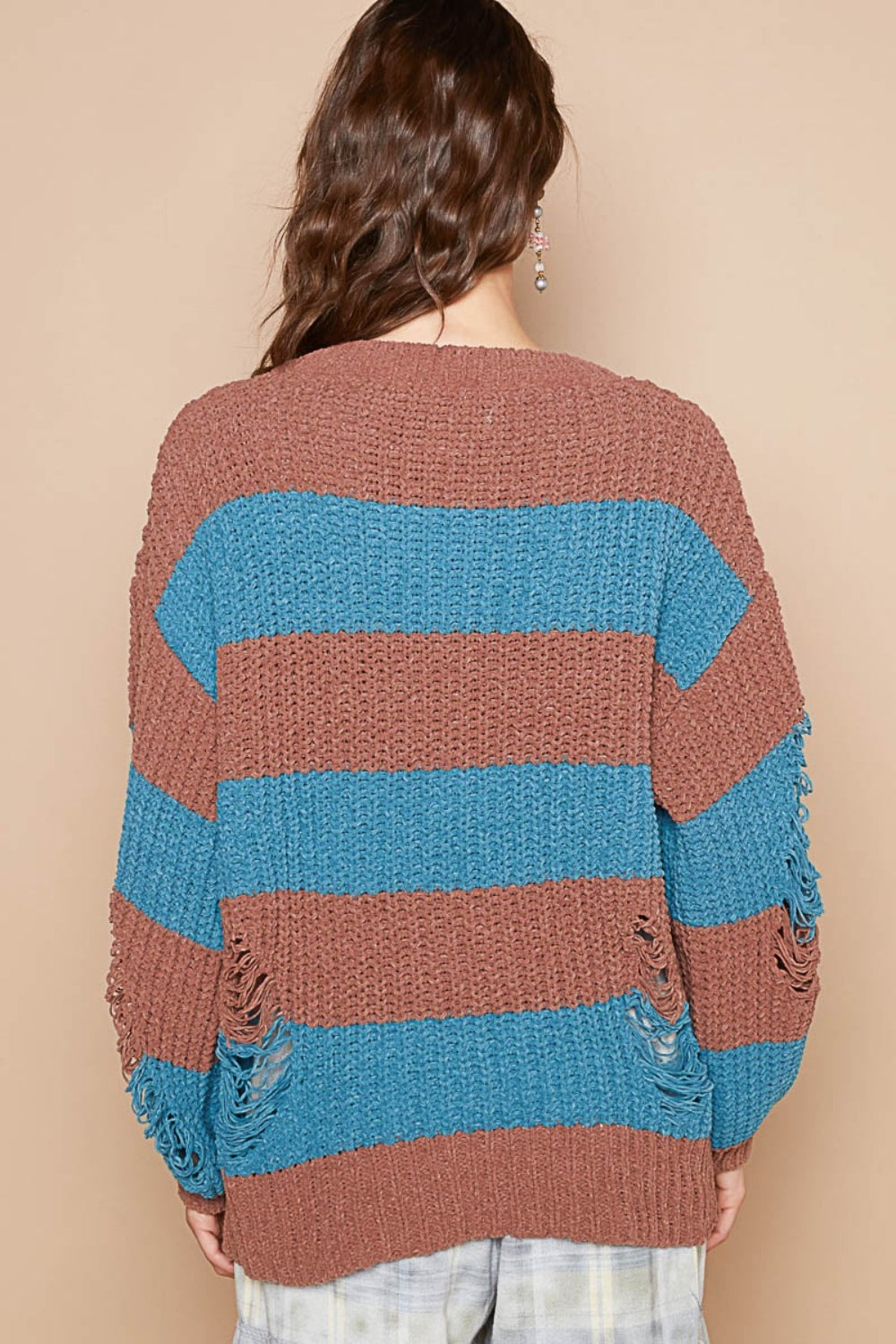 A person with wavy hair is wearing a POL Striped Distressed Long Sleeve Sweater in brown and blue chenille material, paired with light-colored pants, standing against a plain beige background.