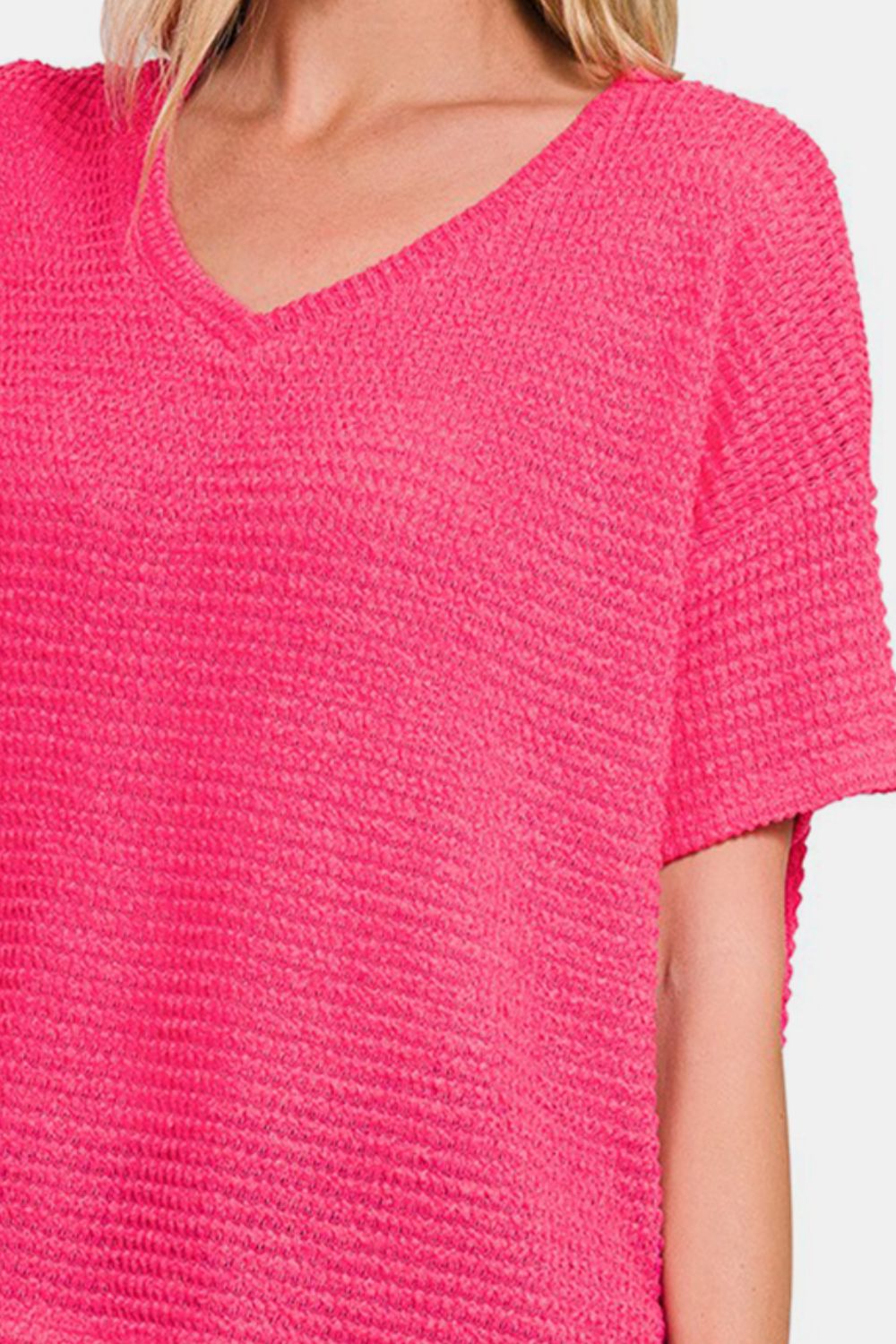 A woman wearing a loose-fitting, bright pink Zenana Drop Shoulder Short Sleeve Jacquard Knit Top with a V-neck and blue jeans touches her hair with one hand and looks at the camera.