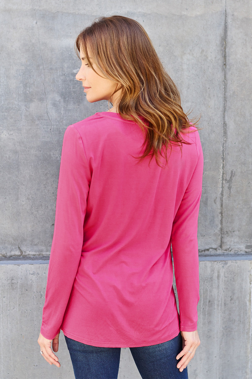 A woman with light brown hair stands against a concrete wall, wearing the Basic Bae Full Size V-Neck Long Sleeve Top in olive green and blue jeans, with a white backpack on her shoulder. Her outfit exudes basic style and is made from polyester, ensuring it’s both comfortable and suitable for machine wash cold. She is looking at the camera.