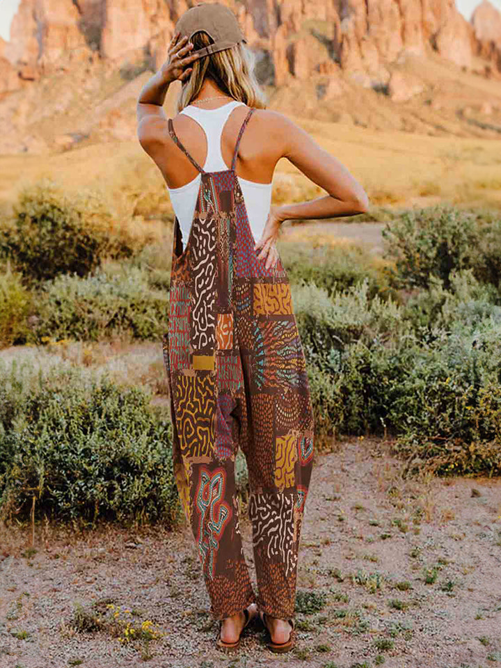 A person standing outdoors in front of rocky terrain, wearing a Full Size Printed V-Neck Sleeveless Jumpsuit and a hat.