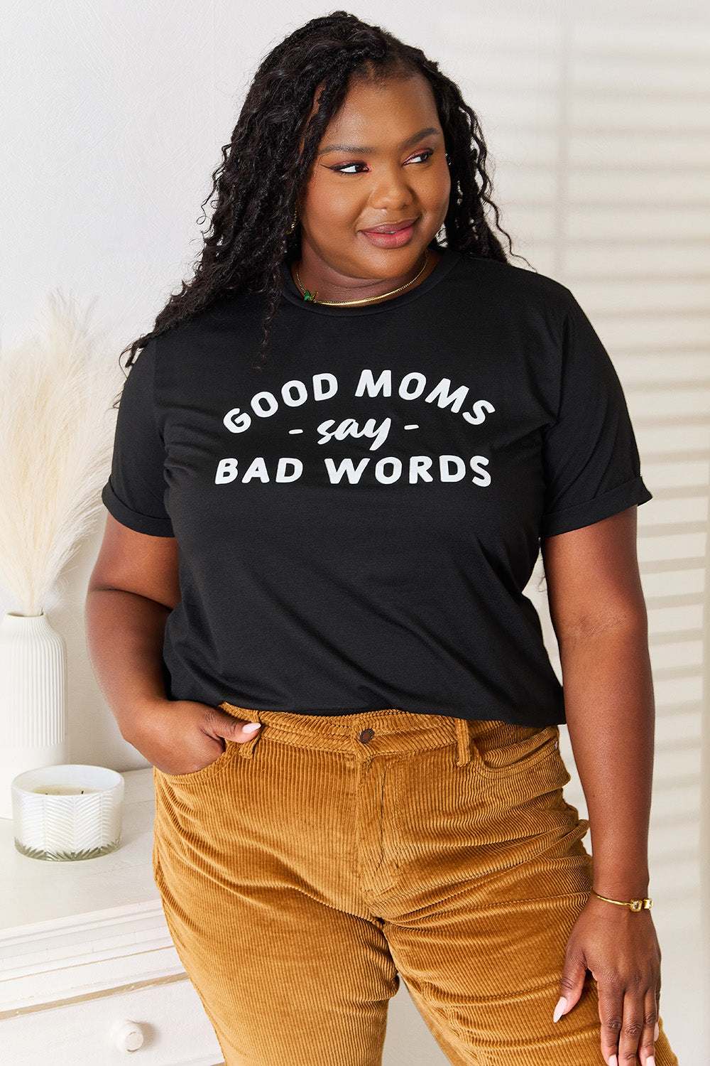 A woman stands in a room with a white wall, where light filters through the blinds, subtly showcasing her rebellious side while wearing the Simply Love GOOD MOMS SAY BAD WORDS Graphic Tee.