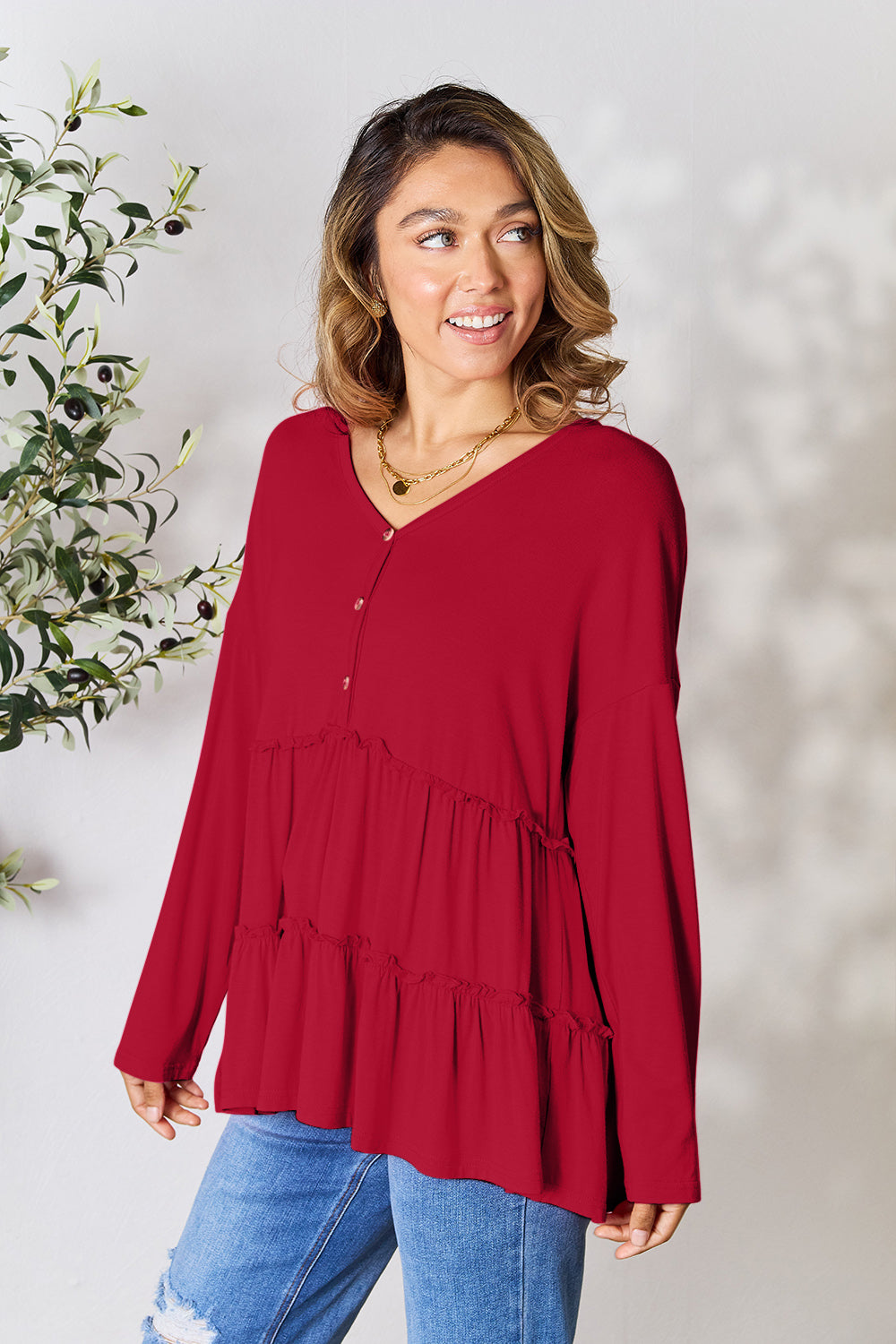 Person stands indoors near a plant, wearing the Double Take Half Button Long Sleeve Ruffle Hem Blouse in gray and blue jeans, smiling while looking to the side. The slightly stretchy fabric of the blouse adds comfort while its buttoned design offers a touch of elegance.