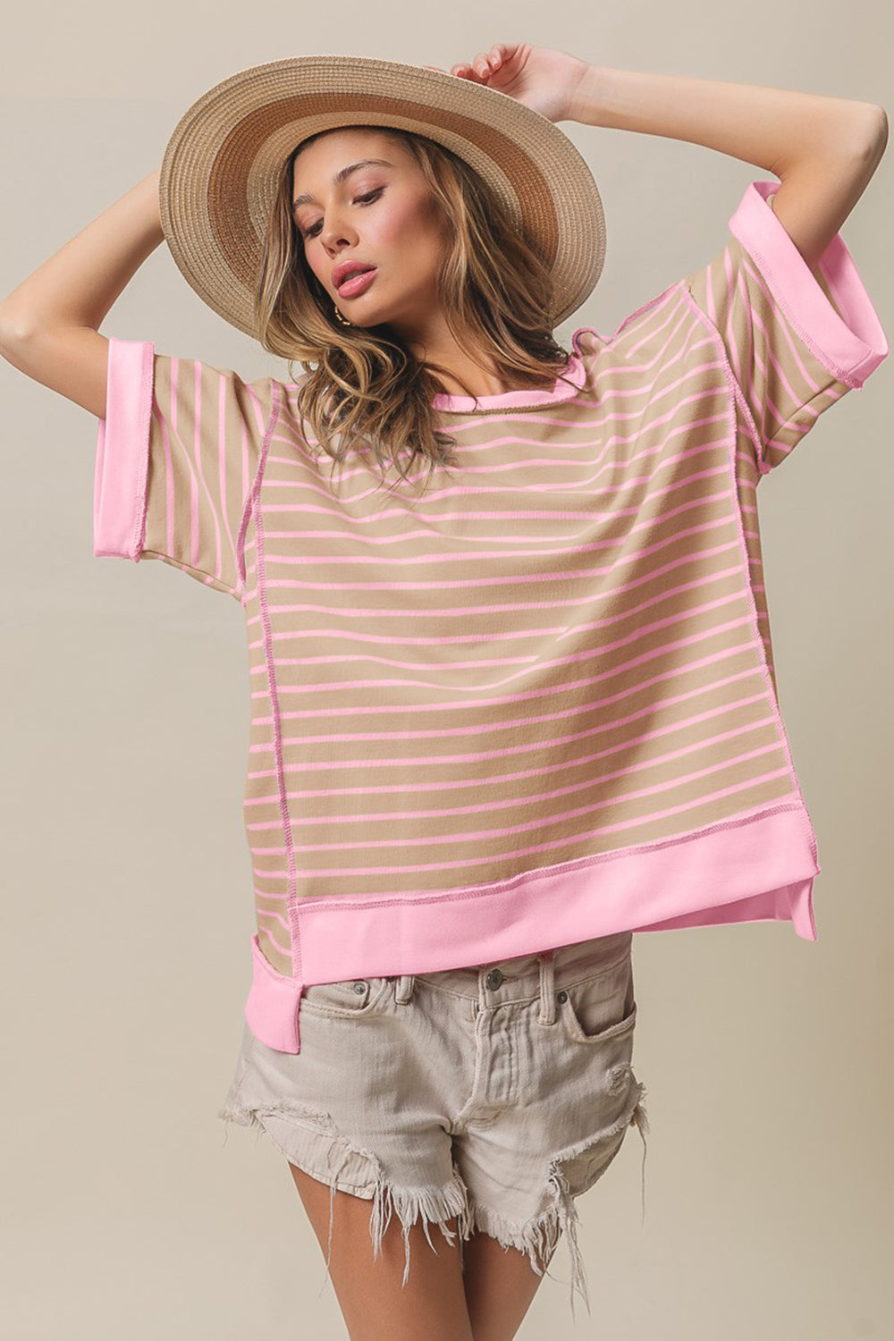 A person wearing a wide-brimmed hat, an oversized BiBi Exposed Seam Stripe Contrast T-Shirt, and distressed denim shorts stands with one arm raised and eyes closed against a beige background.