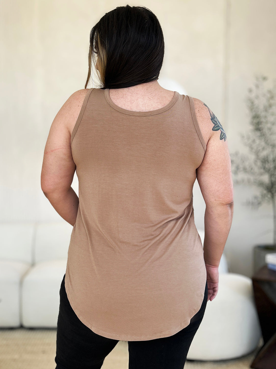 A woman wearing sunglasses, blue jeans, and the Basic Bae Full Size Round Neck Curved Hem Tank is standing in front of a concrete wall, holding her glasses with one hand and carrying a beige shoulder bag. Her basic style complements the slightly stretchy fabric of her outfit, ensuring comfort while maintaining chic simplicity.