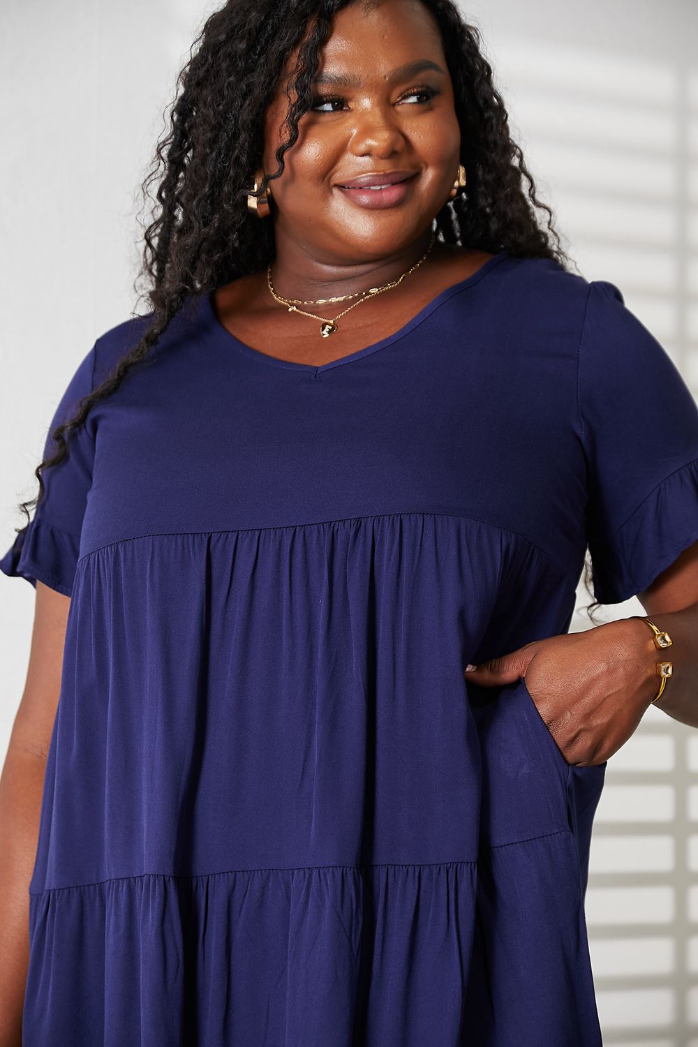 A woman stands indoors wearing the Mandy V-Neck Flounce Sleeve Tiered Dress in navy blue. She has one hand on her hip and is smiling.