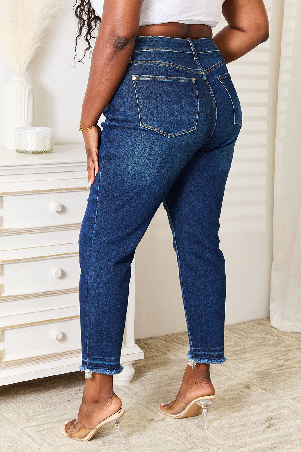 A person wearing Judy Blue Full Size High Waist Released Hem Slit Jeans in indigo wash and a white cropped top stands by a white dresser, showcasing a flattering fit.