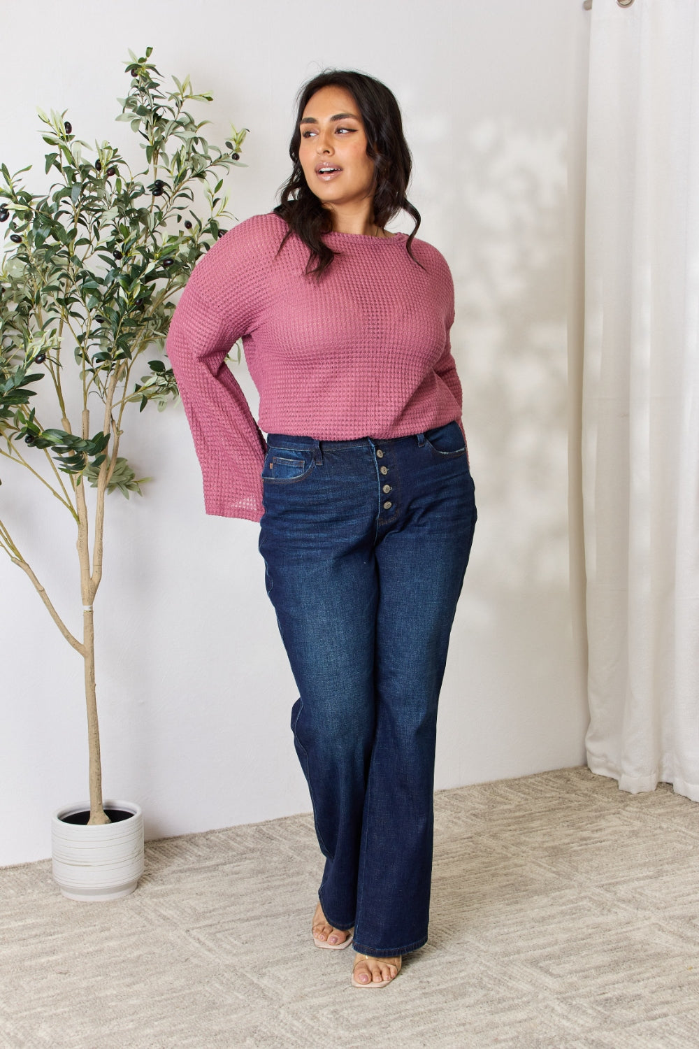 A person wearing a Culture Code Full Size Waffle-Knit Round Neck Long Sleeve Blouse in pink and jeans stands in front of a light background, complemented by a leafy plant on the left.